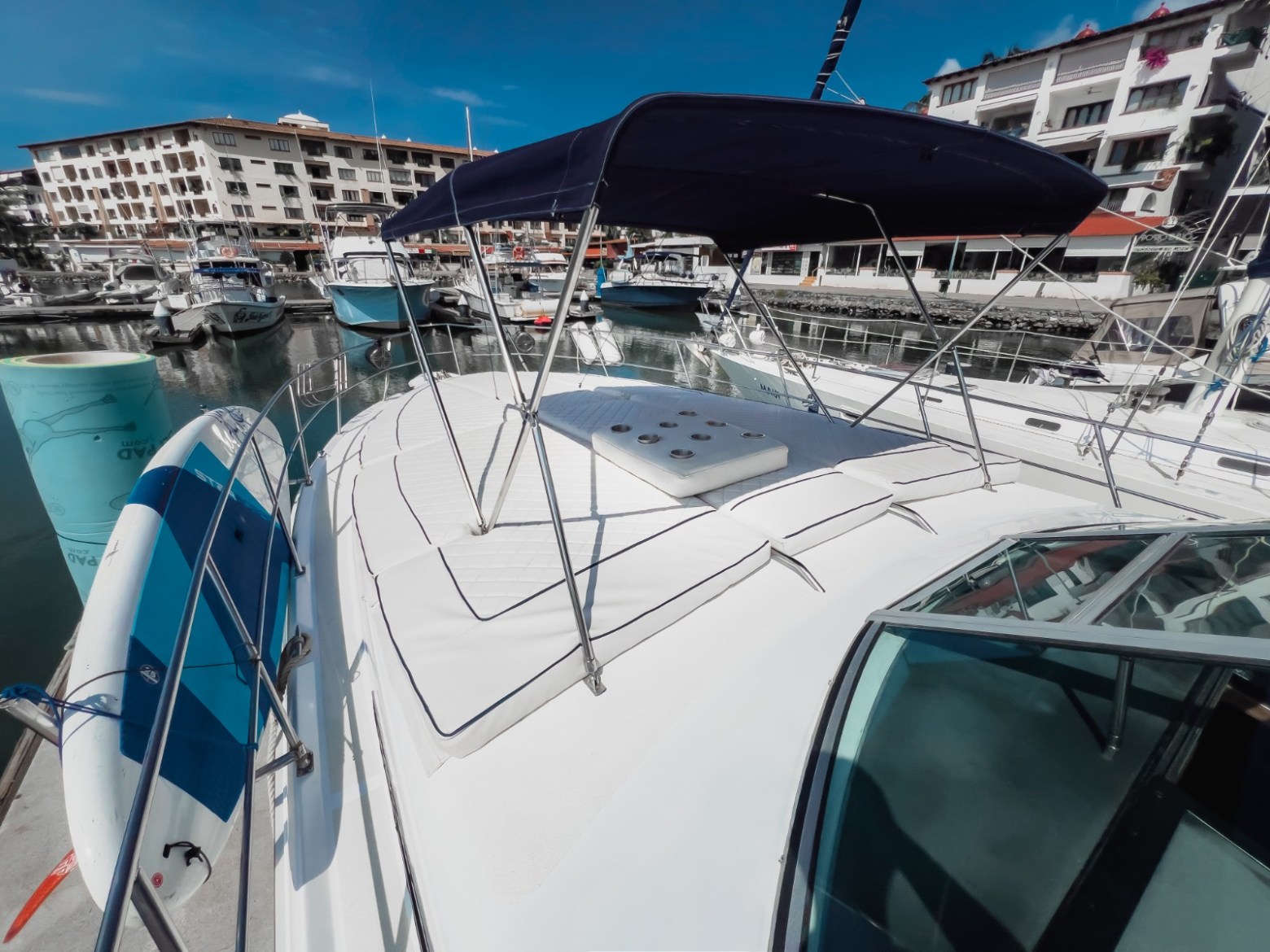 View of a yacht's deck with a blue canopy, docked in a marina with several boats and buildings in the background.