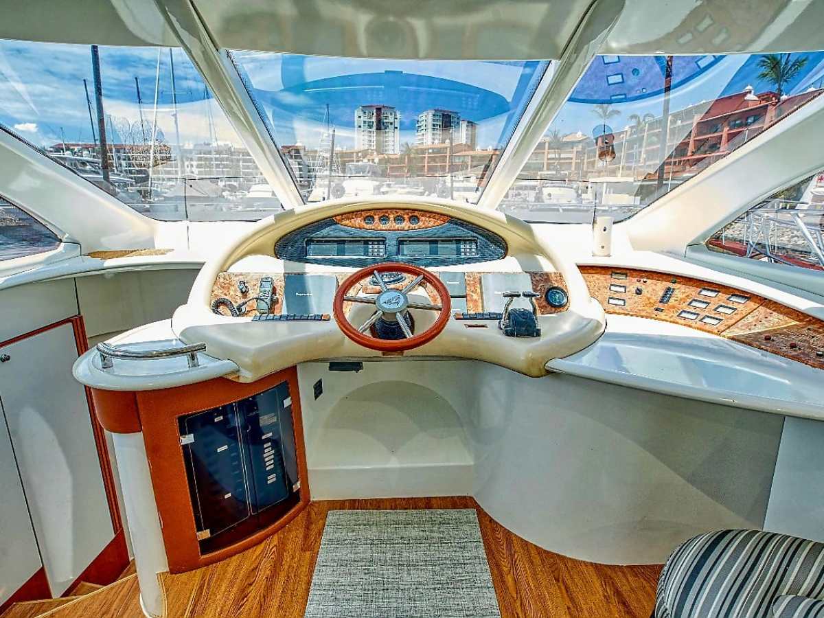 Boat cockpit with steering wheel, gauges, and marina view through windows.