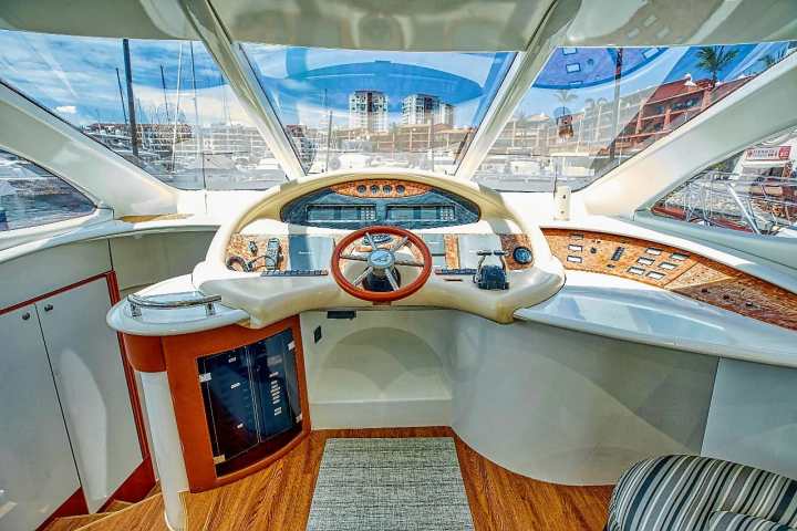 Boat cockpit with steering wheel, gauges, and marina view through windows.
