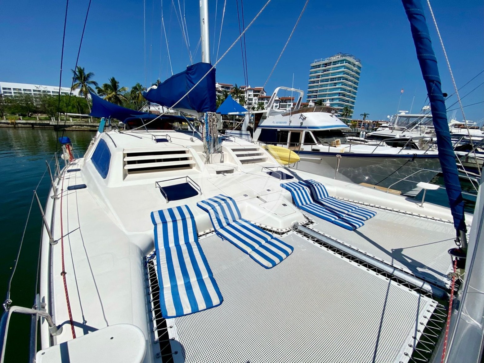 Sailboat deck with blue-striped lounge chairs docked near other boats and buildings.