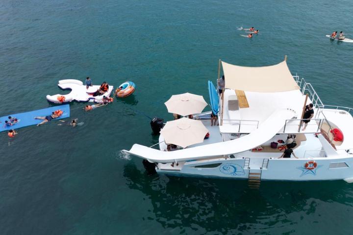 Boat with water slide and people swimming on floating mats in the ocean.