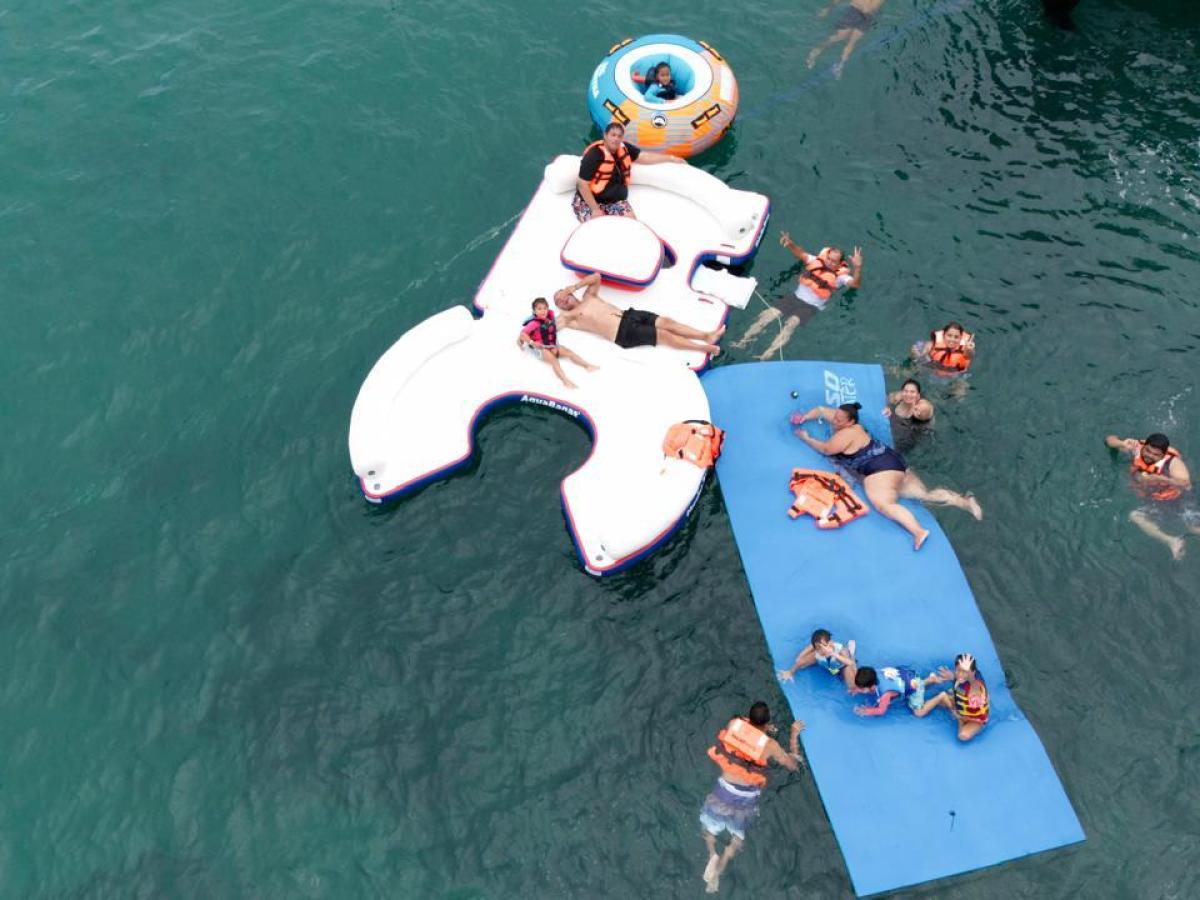 People in life jackets enjoying inflatables and swimming in clear blue water.