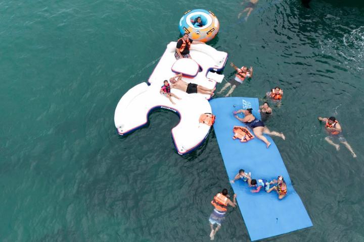 People in life jackets enjoying inflatables and swimming in clear blue water.