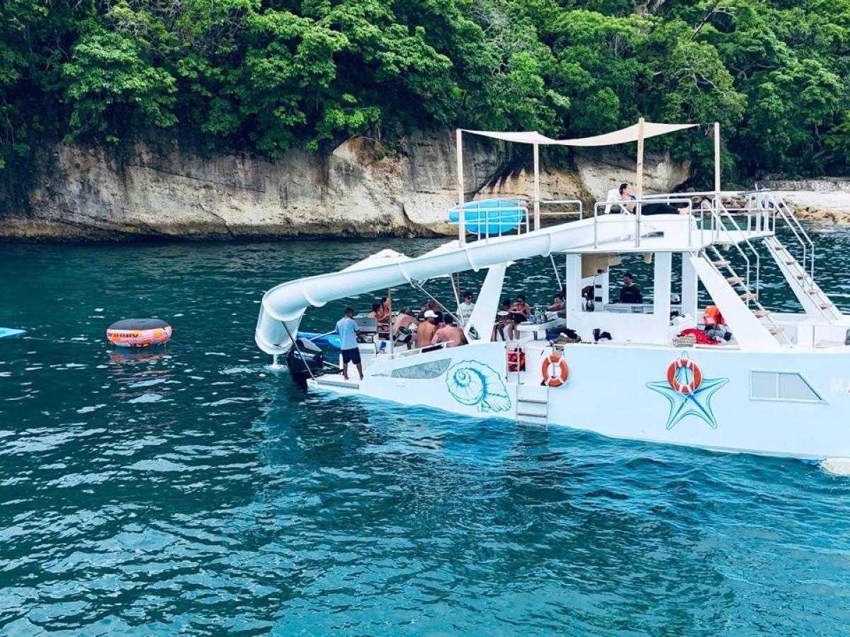 A boat with a slide anchored near a rocky shore and trees, with a floating ring and mat in the water.