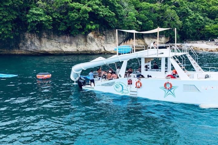 A boat with a slide anchored near a rocky shore and trees, with a floating ring and mat in the water.