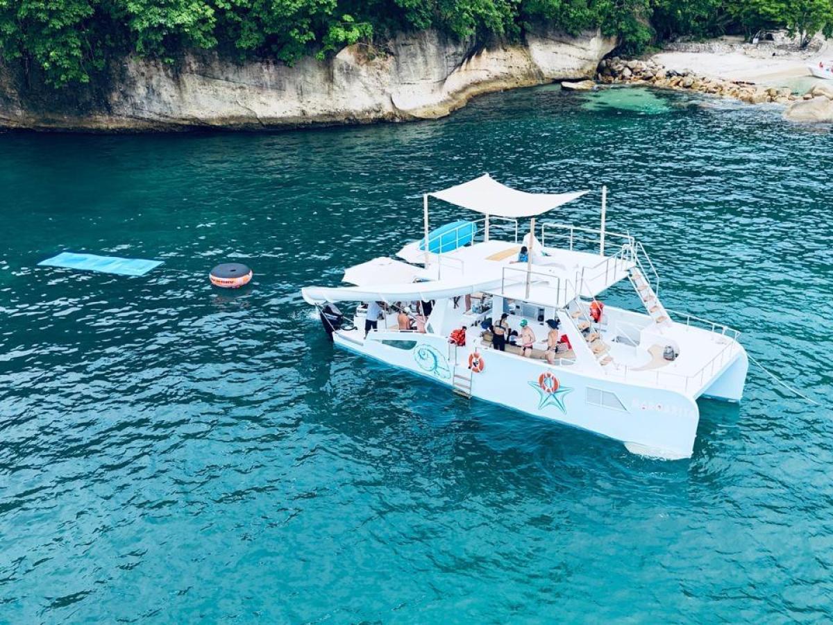 Catamaran with passengers in blue water near rocky coastline with trees.