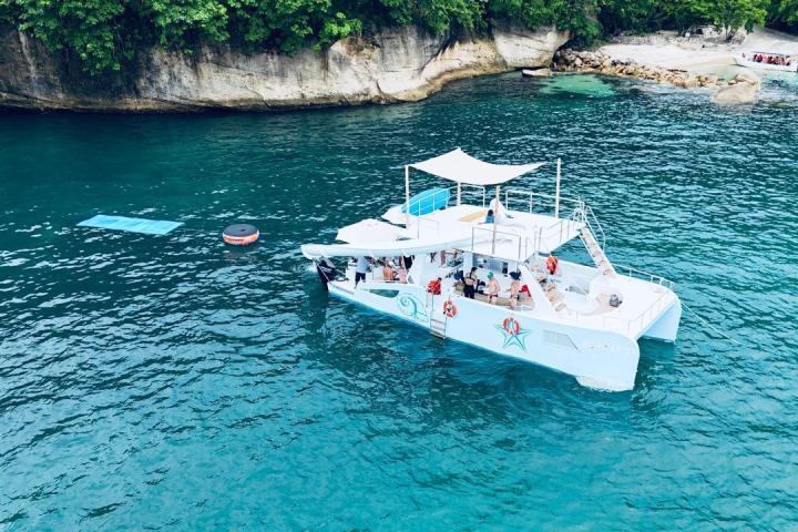 Catamaran with passengers in blue water near rocky coastline with trees.