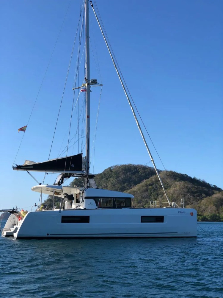 White catamaran with a black sail in calm water near a wooded hillside.