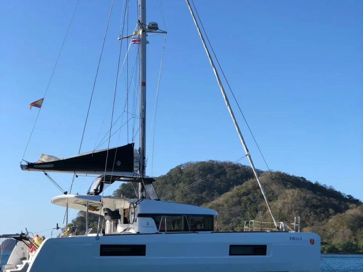 White catamaran with a black sail in calm water near a wooded hillside.