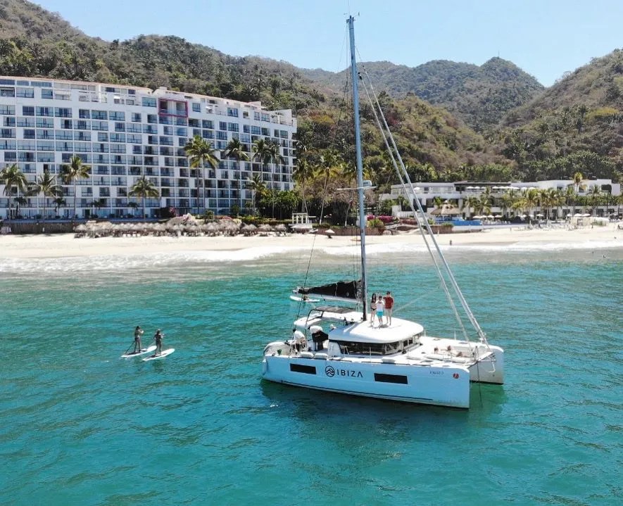 Sailboat near beach with people paddleboarding and hotel in background.