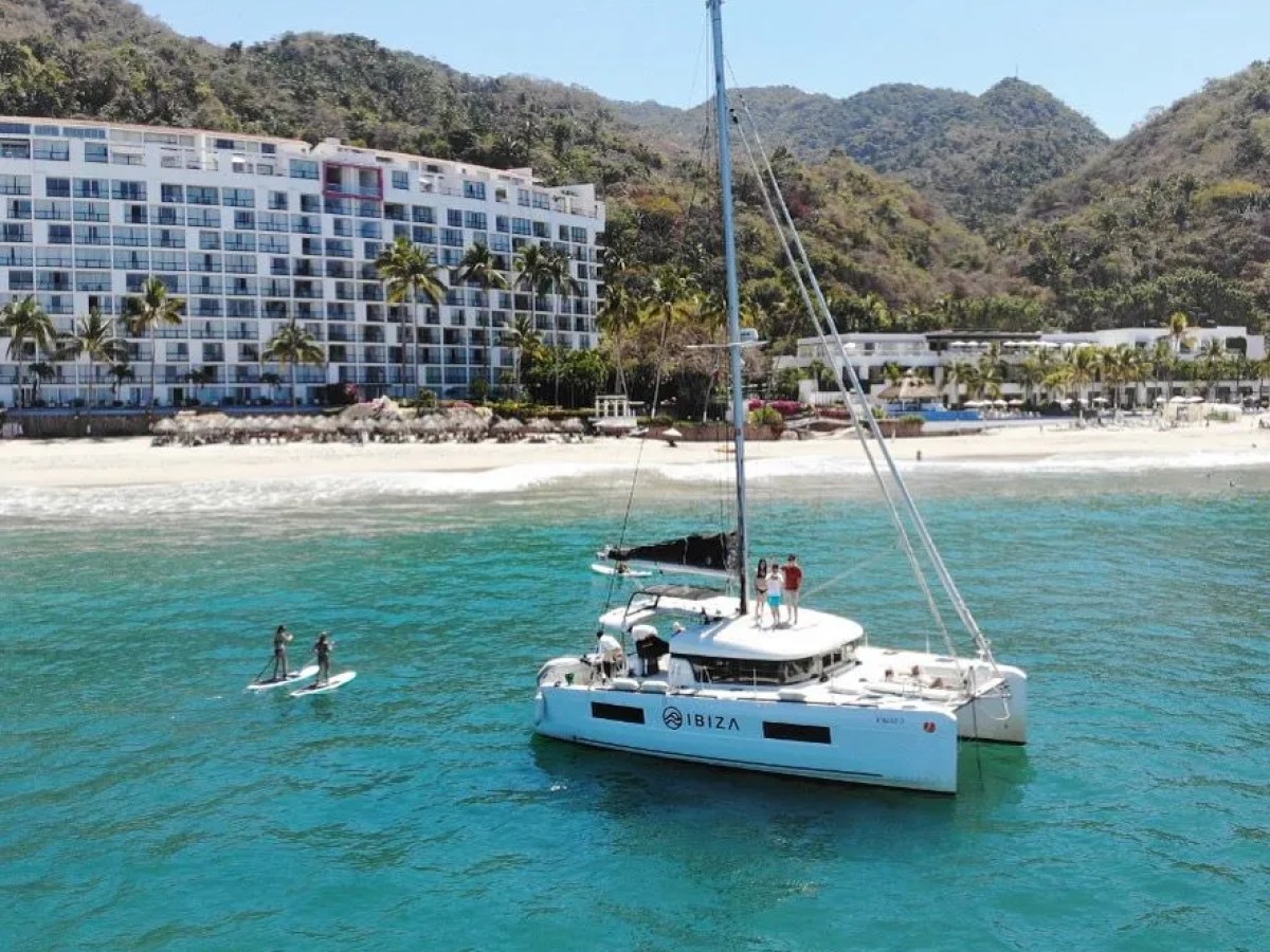 Sailboat near beach with people paddleboarding and hotel in background.