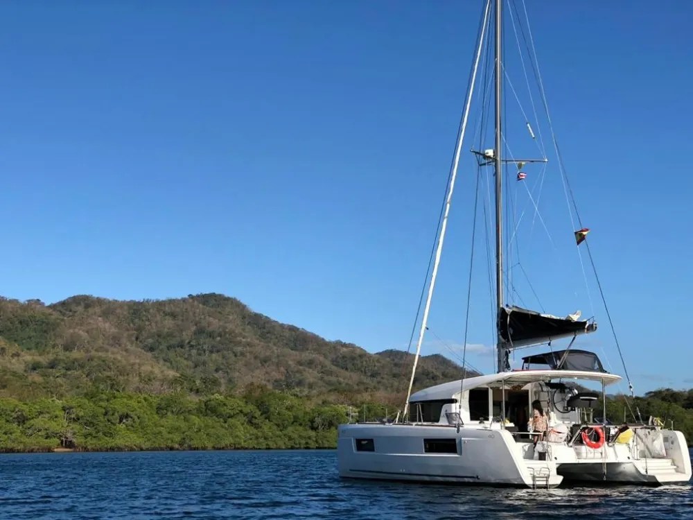A catamaran sailboat anchored on a calm sea with a lush, hilly landscape in the background.