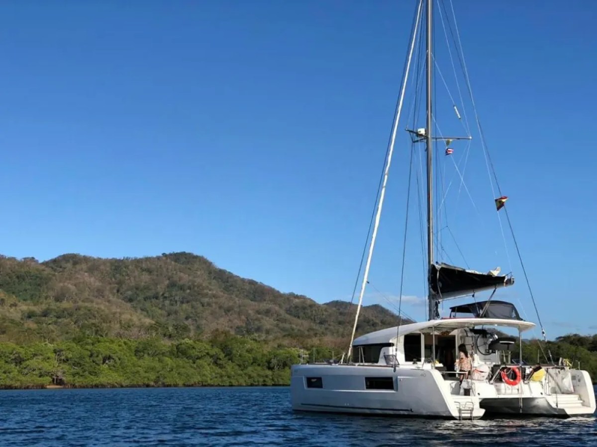 A catamaran sailboat anchored on a calm sea with a lush, hilly landscape in the background.