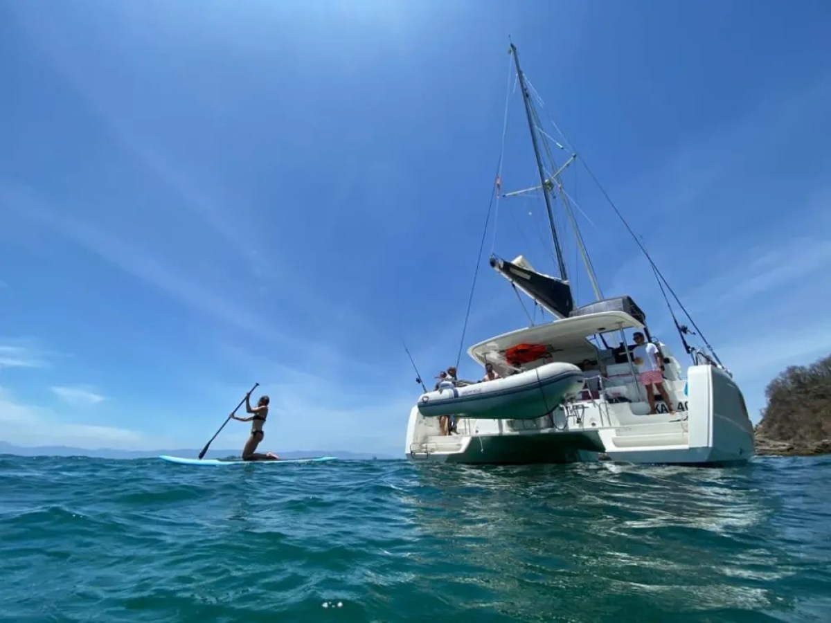 Woman paddleboarding near a catamaran with clear blue sky and ocean.