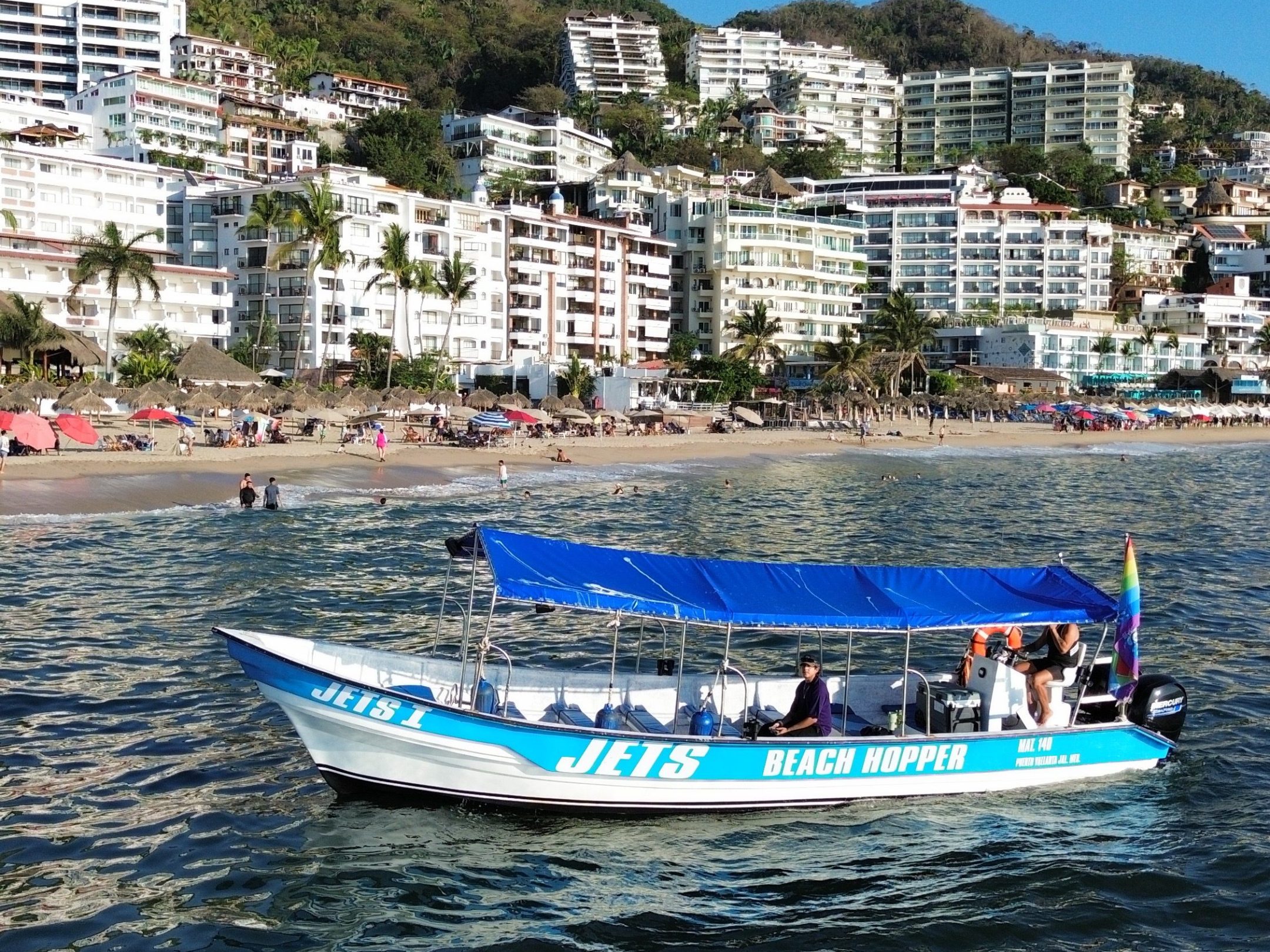 Boat with blue canopy on water near hillside buildings.