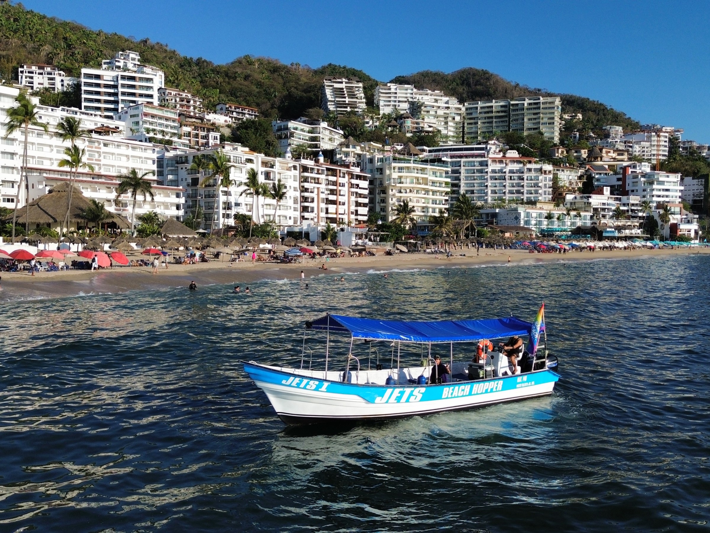 Small boat with blue canopy on ocean near a busy beach and hillside buildings.