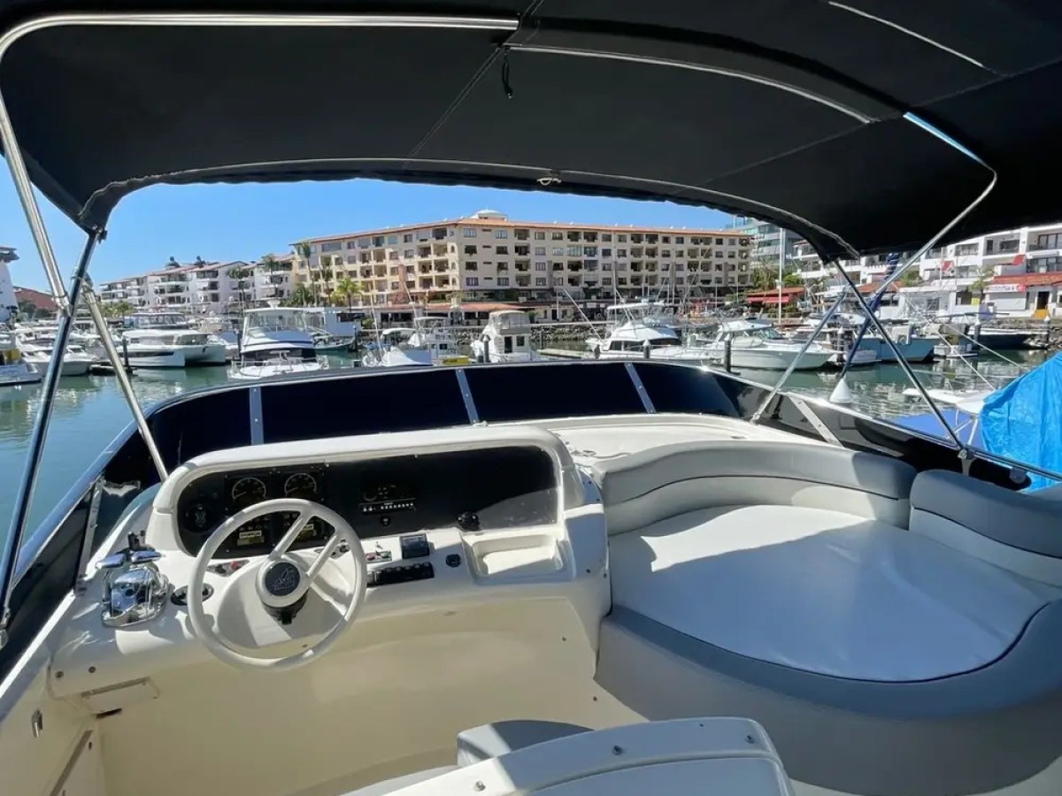 Interior of a boat with a steering wheel, overlooking a marina with moored boats and buildings.