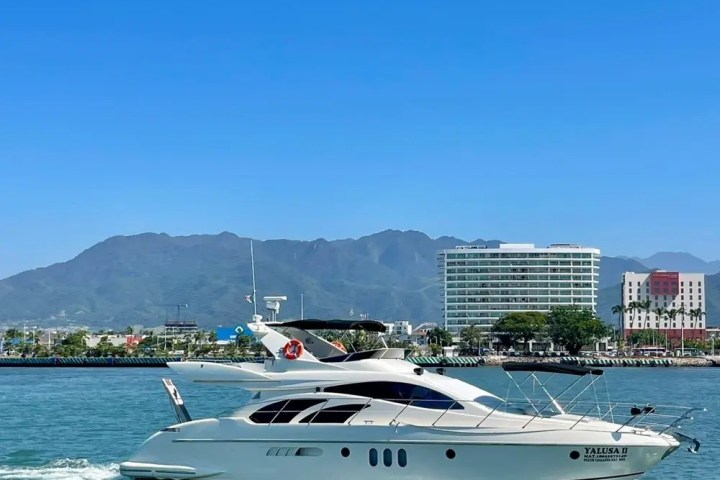 White yacht on water with city and mountains in background under clear blue sky.