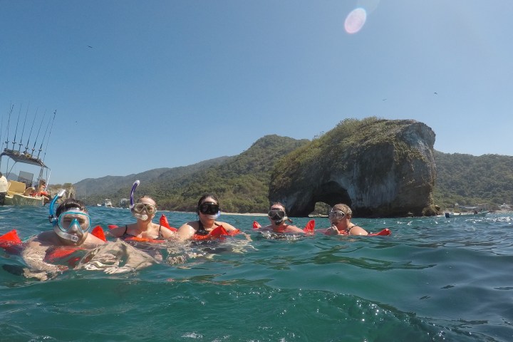Five snorkelers in the ocean near a rocky island under a clear blue sky.