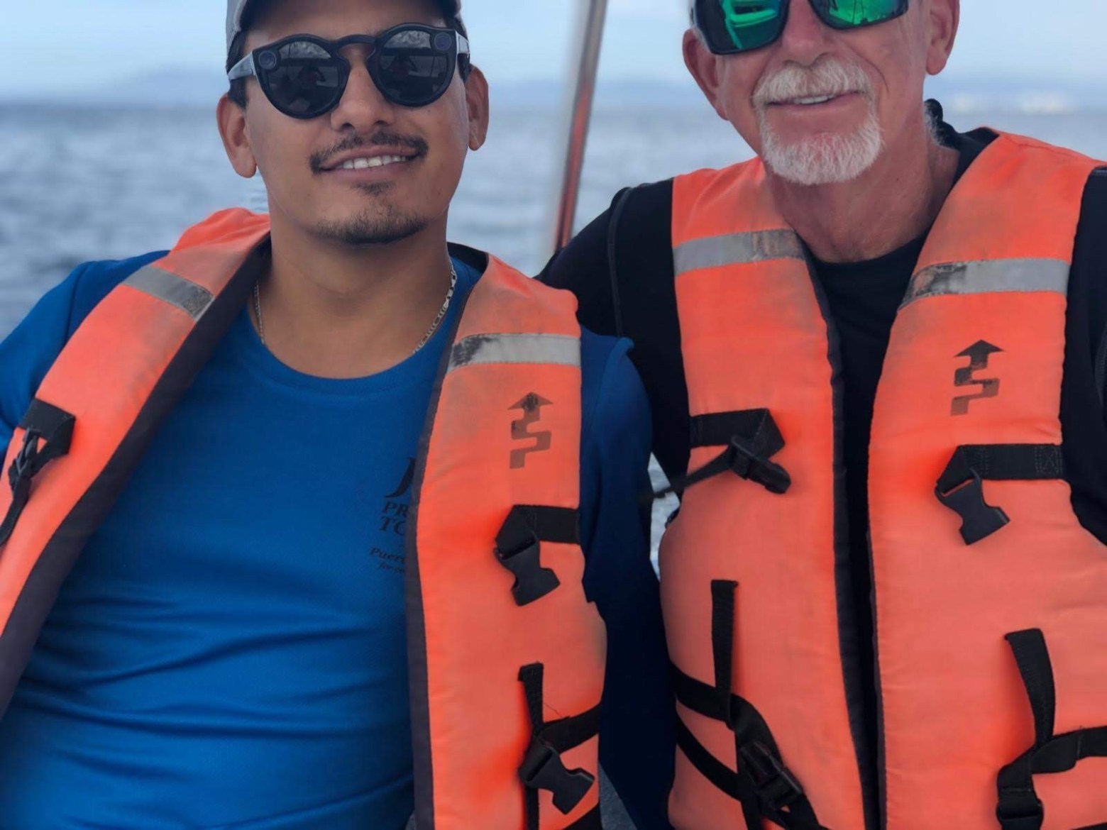 Two men wearing orange life vests on a boat with water in the background.